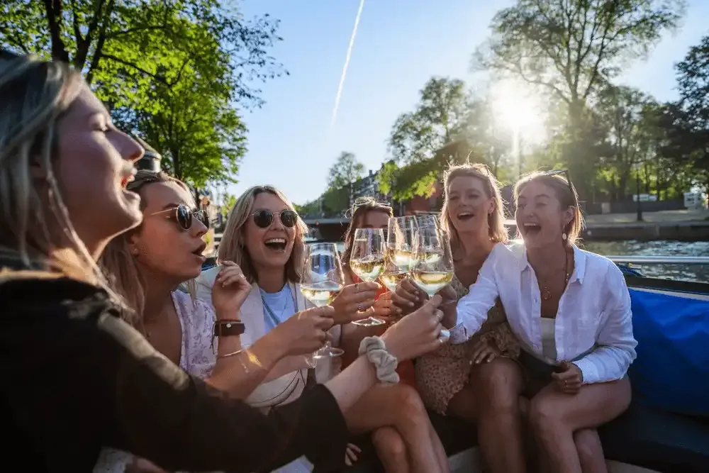 Group of laughing women raising glasses of white wine on a boat in sunlit Amsterdam canal