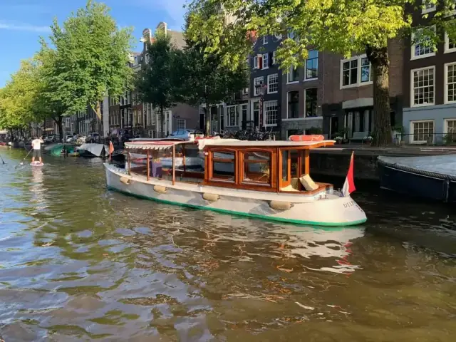 Small wooden-cabin canal boat with white hull on an Amsterdam canal beside tree-lined brick houses