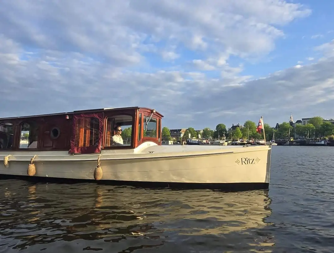 Ritz, a cream-hulled saloon boat with a red wooden cabin and skipper at the helm on Amsterdam water